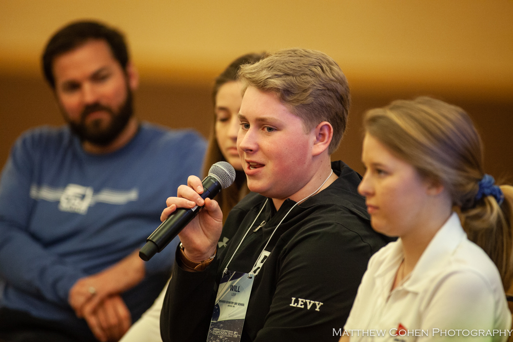 A young person speaks into a microphone during a discussion, with three others sitting beside them and listening attentively. The background is softly blurred.