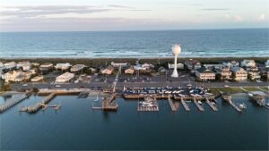 Aerial view of a coastal town with a marina, docked boats, houses, a parking lot, and a large water tower near the shoreline, with the ocean and waves in the background under a partly cloudy sky.
