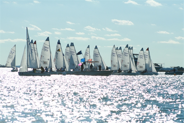 A group of sailboats with white sails gathers on a sparkling, sunlit body of water under a partly cloudy sky, possibly preparing for a race.