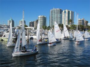 Small sailboats with people in life jackets are racing on a marina, with modern high-rise buildings and palm trees in the background under a clear blue sky.