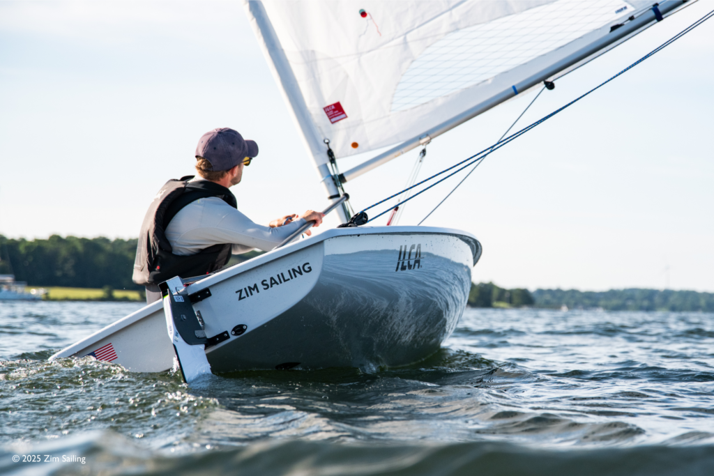 A person wearing a cap and life jacket is sailing a small Zim Sailing dinghy on a lake, leaning to balance the boat as it cuts through the water on a sunny day.