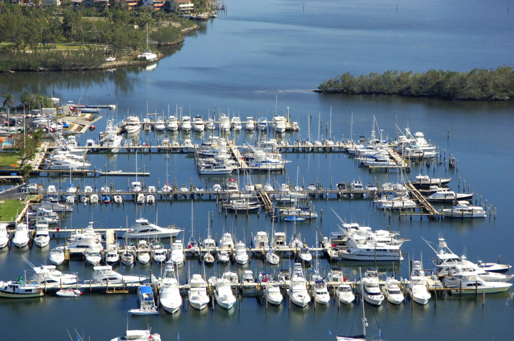 Aerial view of a marina with numerous white boats and yachts docked in rows, surrounded by calm blue water and greenery along the shoreline.