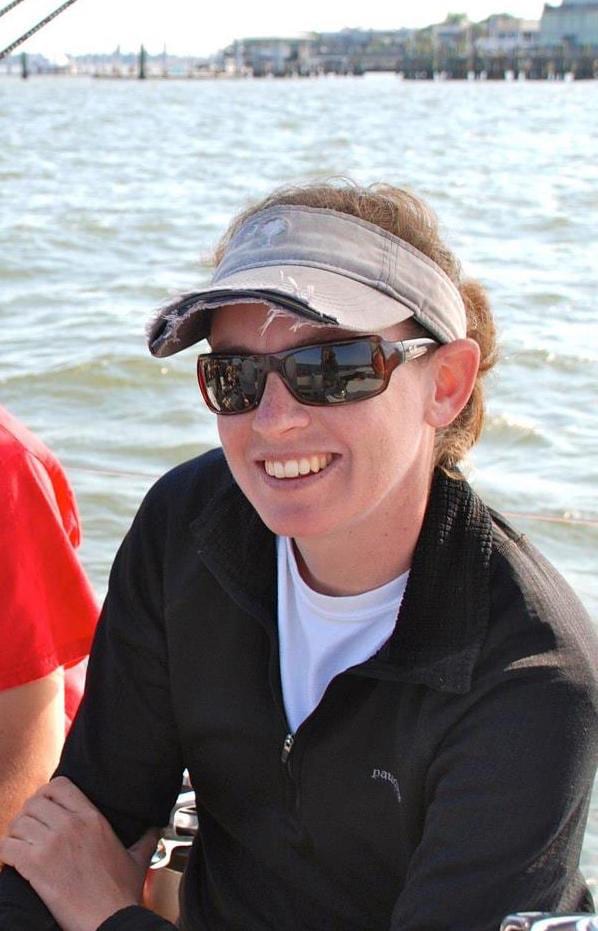 A person wearing sunglasses, a visor, and a black jacket smiles while sitting on a boat with water and buildings in the background on a sunny day.