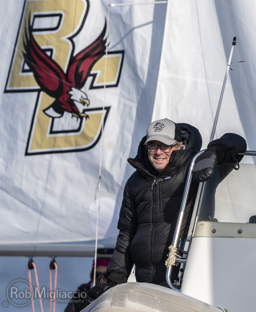 A person in a black jacket and BC hat smiles while standing on a boat, with a large Boston College sail featuring an eagle visible in the background.