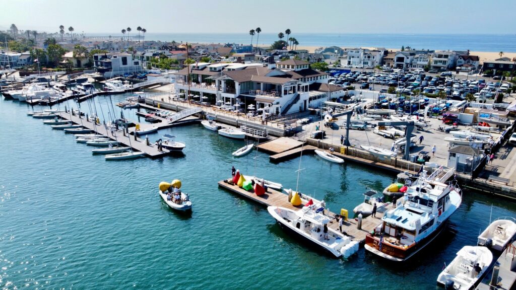 Aerial view of a marina with several docks, boats, and yachts on the water. Buildings, parked cars, and palm trees line the shore, with the ocean and a beach visible in the background.