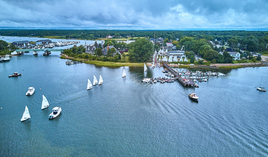 Aerial view of a harbor with sailboats on the water, a dock with boats, and a small town surrounded by trees under a cloudy sky.