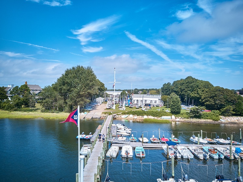 A dock lined with small boats extends over the water toward a marina, with a red flag on a pole in the foreground. Buildings and trees are visible under a blue sky with scattered clouds.