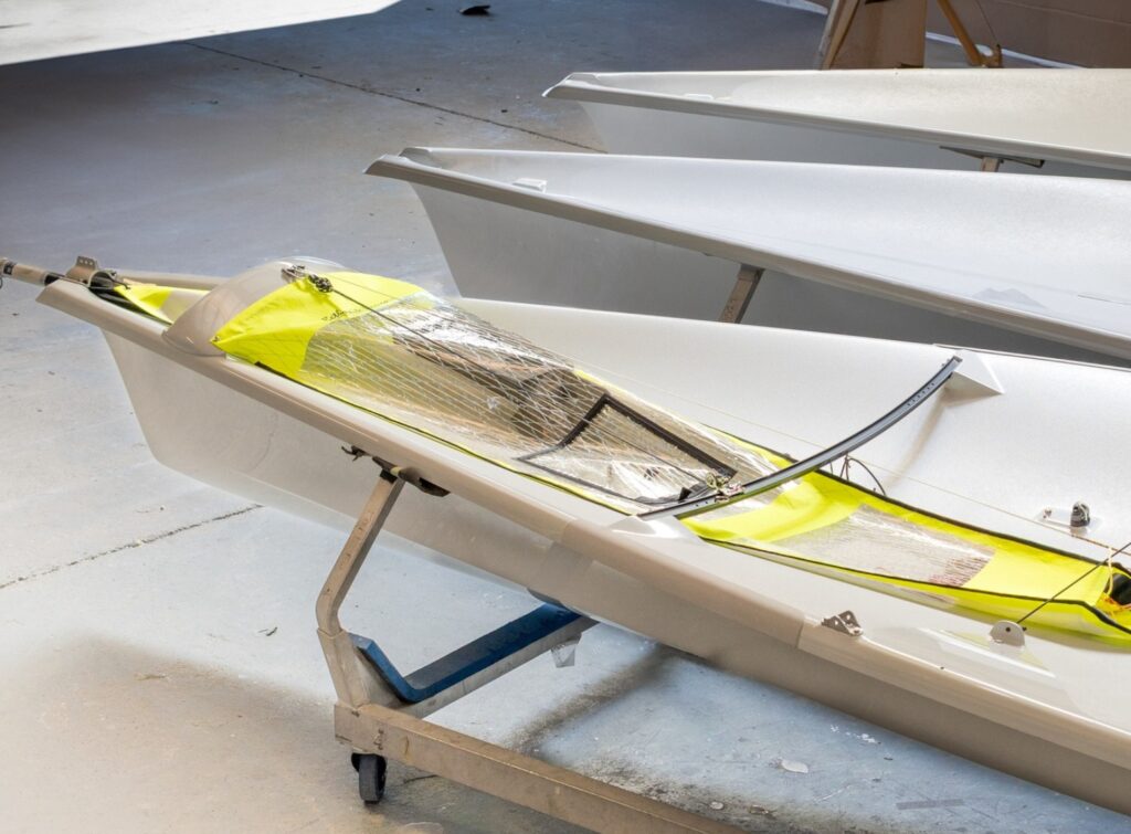 A close-up of a sleek, white racing sailboat with a yellow cockpit, resting on a metal trolley inside a brightly lit workshop or storage area. Other similar boats are lined up nearby.
