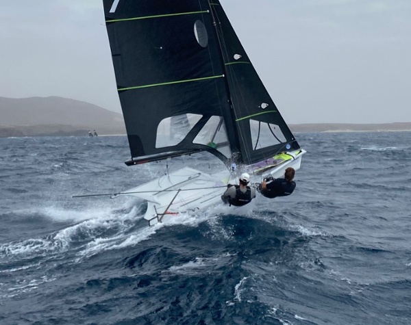 Two sailors are leaning out from a small, fast sailboat with a black sail as it speeds over choppy, dark blue water on a cloudy day. Distant hills and a faint coastline are visible in the background.