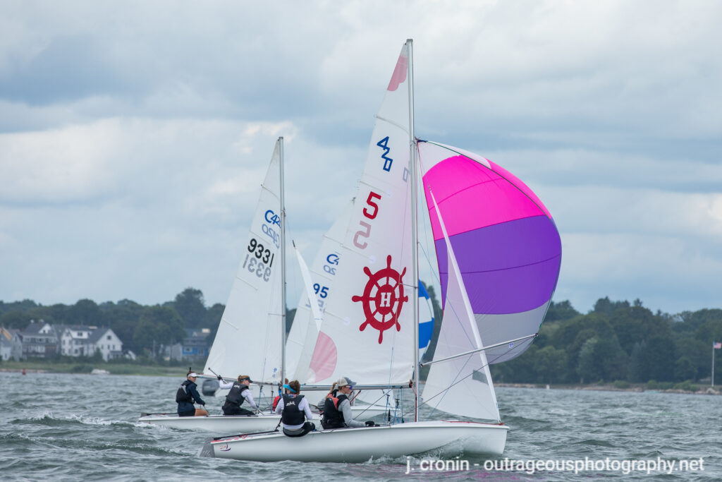 Two sailboats race on the water, with one boat displaying a pink and purple spinnaker and a red ship wheel symbol on its sail. A shoreline with houses and trees is visible in the background under a cloudy sky.