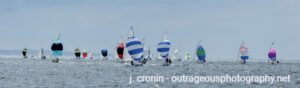 A group of sailboats with colorful spinnakers race across open water under a cloudy sky. The sails display various bright patterns and stripes. Text at the bottom reads “j. cronin - outrageousphotography.net.”.