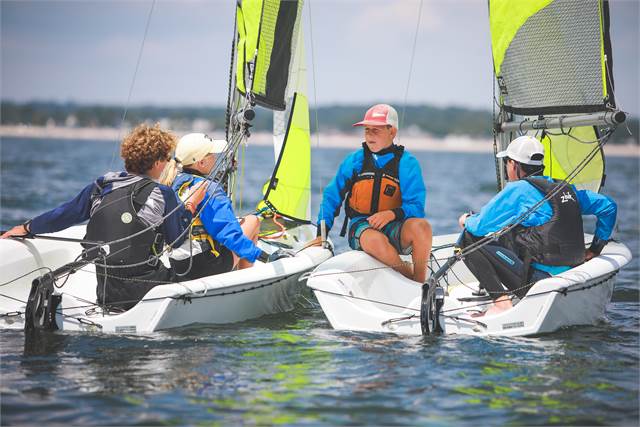 Four people in life jackets sit in two small sailboats on the water, talking to each other. The sailboats have bright yellow and gray sails. The background shows a distant shoreline under a partly cloudy sky.