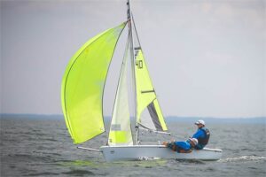 Two people sail a small white dinghy with bright neon yellow sails on open water under a cloudy sky. Both sailors wear blue life jackets and caps as they lean back to balance the boat.