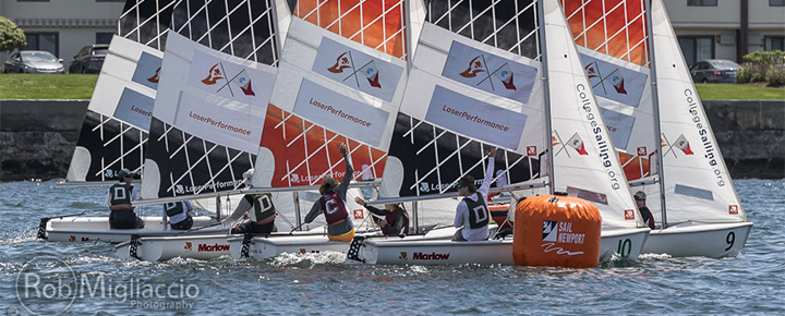 Sailboats with crews compete closely around an orange buoy marked 'Sail Newport' on a sunny day, with apartment buildings visible in the background. Each boat displays sponsor banners on its sails.