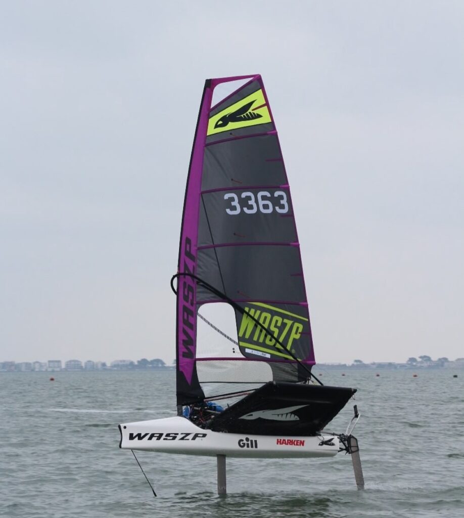 A person sails a WASZP hydrofoil dinghy with sail number 3363 over water, lifting above the surface. The sail is black and purple with yellow and green accents. The background shows a distant shoreline under a cloudy sky.