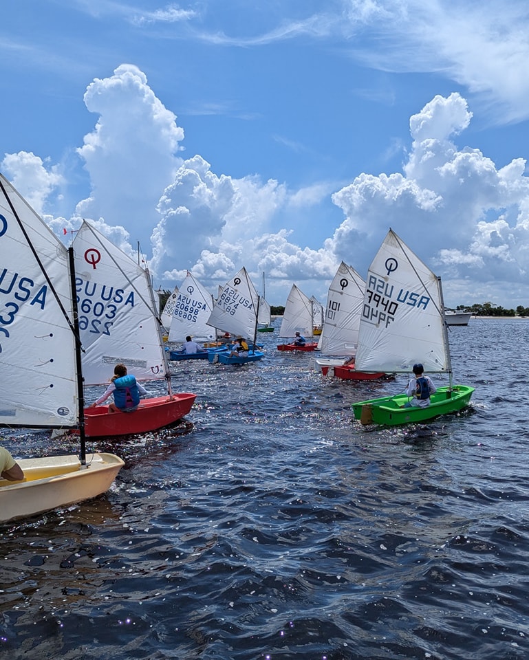 Several small sailboats with young sailors are racing on a sunny day, with blue sky and large puffy clouds in the background. The sails display numbers and 'USA' markings. The water is dark and reflective.