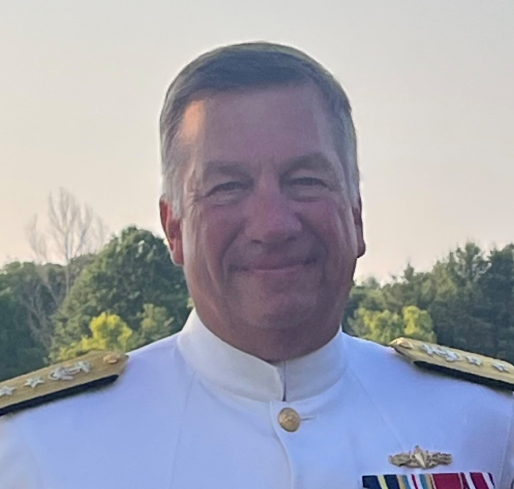 A man in a white military dress uniform with gold epaulets and medals stands outdoors, smiling, with trees and a clear sky in the background.