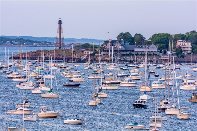 Numerous sailboats and motorboats float in a busy harbor near a rocky shoreline with a tall lighthouse and several large coastal houses surrounded by greenery in the background.
