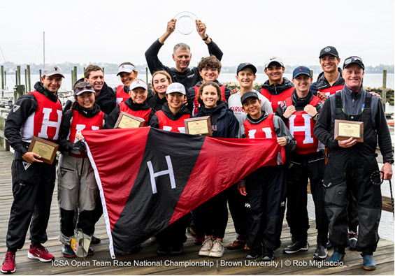 A group of 14 sailors in matching Harvard gear pose on a dock, smiling and holding plaques and a Harvard flag after winning the ICSA Open Team Race National Championship. One person holds a glass trophy above his head.