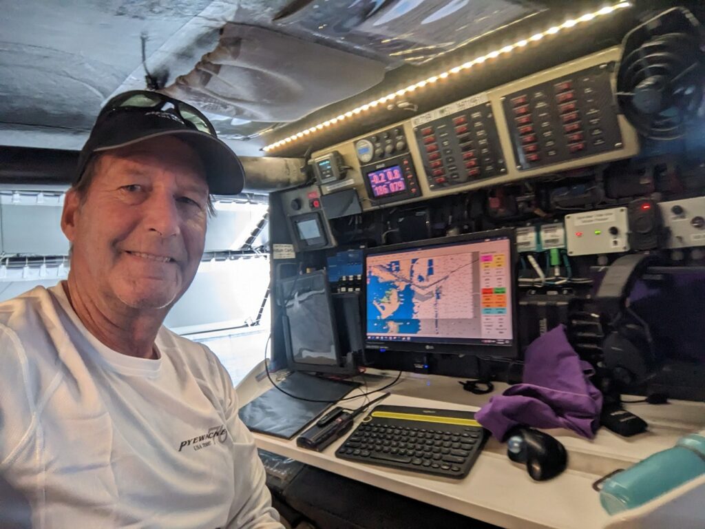 A man smiles at the camera while sitting at a desk with navigation equipment, monitors, and controls, possibly inside a boat or a cockpit. He wears a white shirt and a black cap, and sunlight shines from above.
