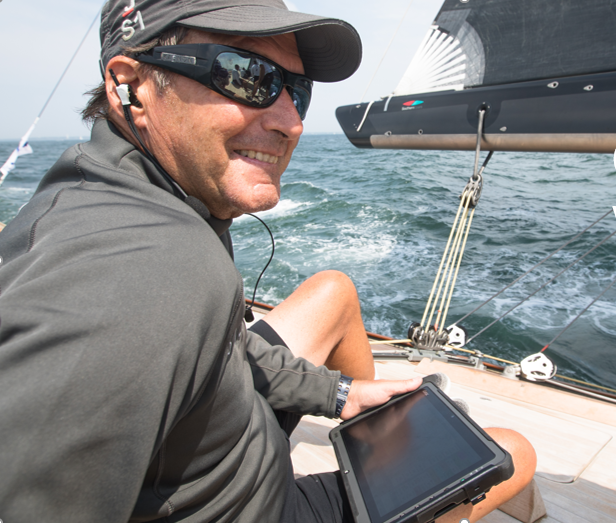 A man wearing a cap, sunglasses, and headphones smiles while sitting on a sailboat holding a tablet, with the sea and sails visible in the background.