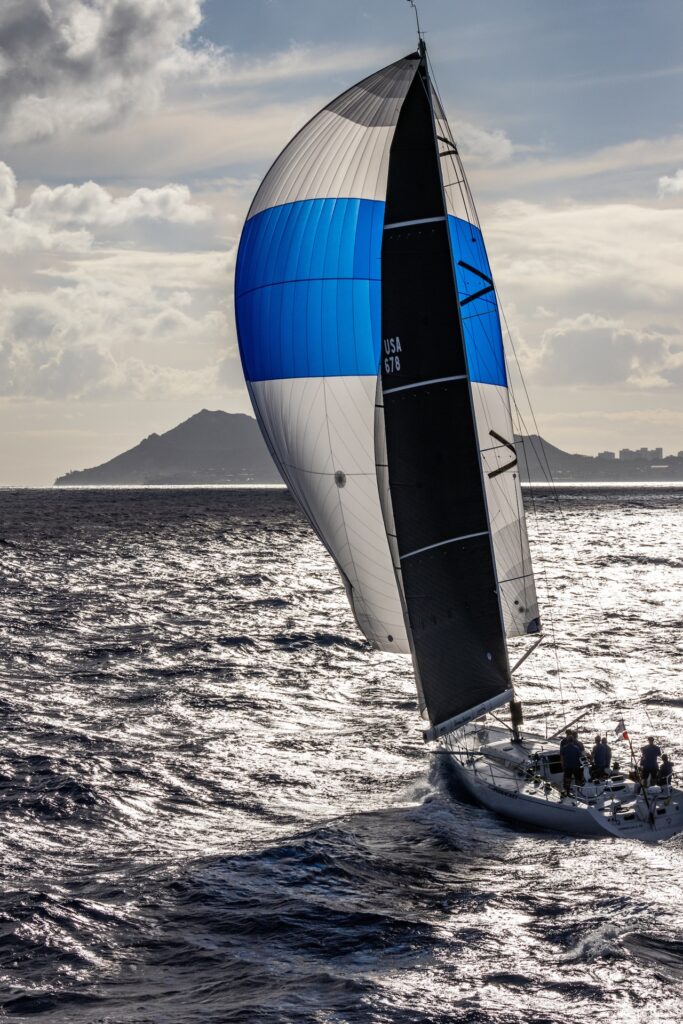 A sailboat with a blue, white, and black spinnaker sails across a sunlit ocean with an island and mountains visible in the background under a partly cloudy sky.