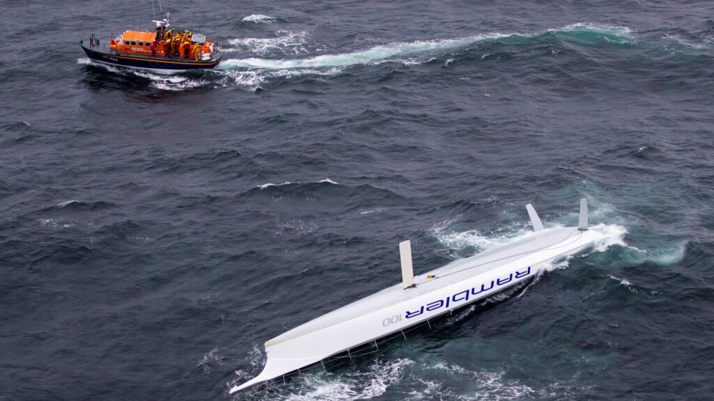 A lifeboat sails near an upturned vessel labeled "RAMBLER 100" in rough, dark ocean waters. The scene suggests a rescue operation following the capsizing of the boat.