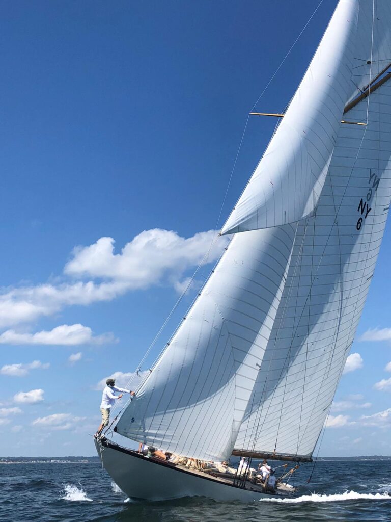 A sailboat with people on board glides across the water on a sunny day. One person stands at the front adjusting a sail, while others sit along the deck. The sky is blue with scattered clouds.