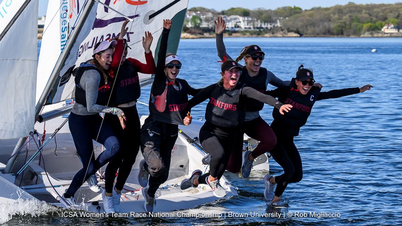 Five women wearing Stanford team gear celebrate energetically on a sailboat, jumping and smiling with joy, while sailing on calm water near a shoreline with trees and houses in the background.
