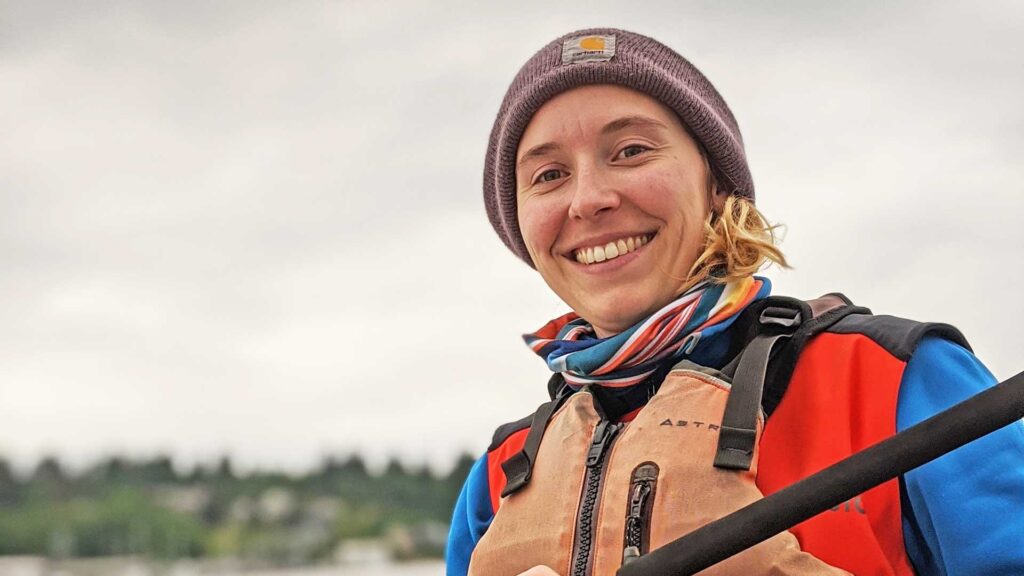 A person wearing a brown beanie, colorful scarf, blue shirt, and orange life jacket smiles while holding a paddle, with an overcast sky and blurred landscape in the background.