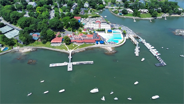 Aerial view of a waterfront marina with docks, swimming pools, tennis courts, several buildings, and boats anchored in the water; surrounded by trees and nearby residential houses.