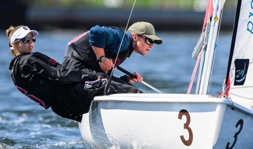 Two sailors wearing sunglasses and sailing gear lean out of a small white sailboat marked with the number 3, gliding quickly over the water during a race.