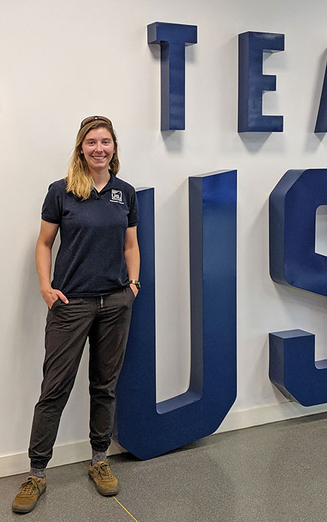 A woman in a navy polo shirt and dark pants stands smiling with one hand in her pocket next to a large wall sign that reads "TEAM USA" in blue letters.