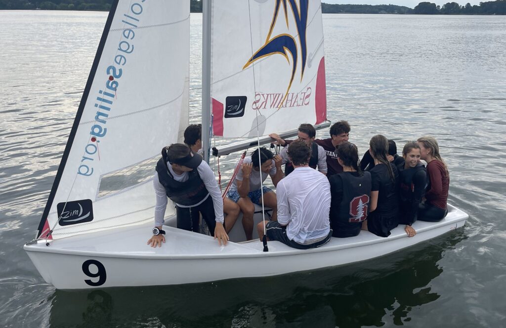 Eleven people in life jackets are crowded together on a small white sailboat with the number 9, floating on calm water with trees in the background.