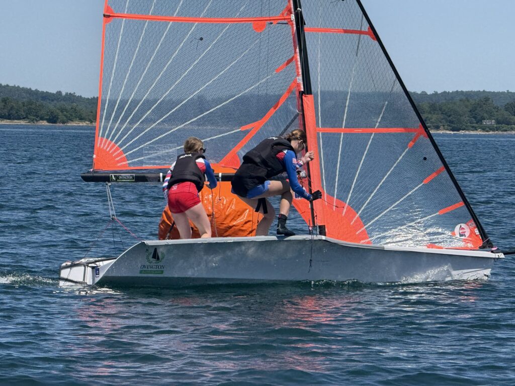 Two people wearing life jackets work together to adjust equipment on a small sailboat with bright orange sails, sailing on a blue lake with forested shoreline visible in the background.