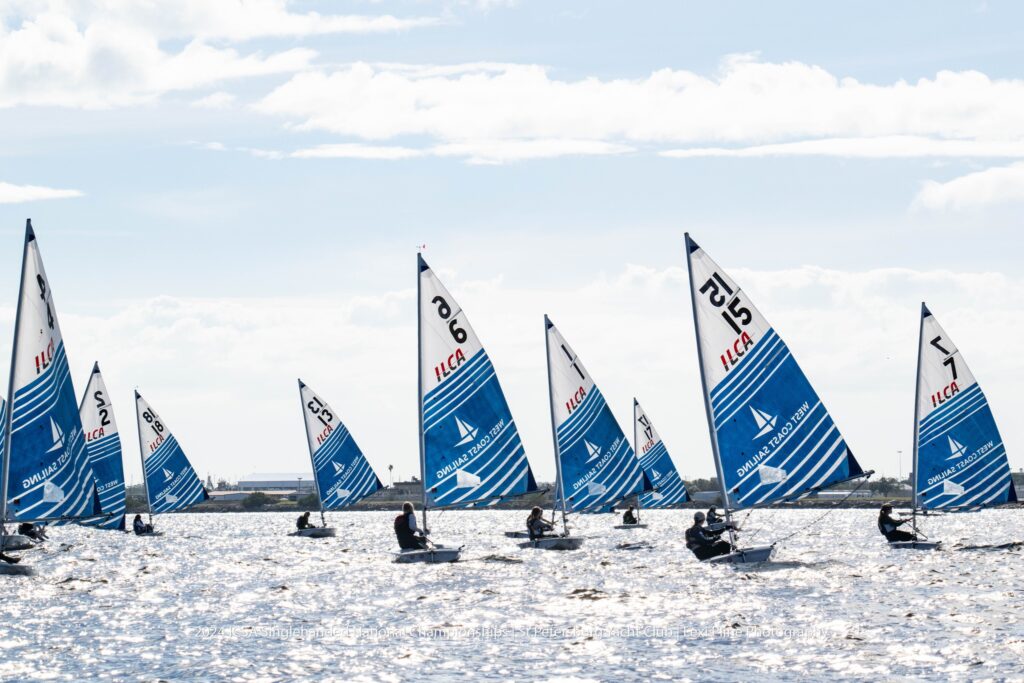 A group of small sailboats with blue and white sails are racing on a sunlit body of water under a partly cloudy sky. The sails display various numbers and the letters 'ILCA.