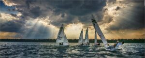 Several small sailboats with people aboard race across a lake under dramatic, cloudy skies with sun rays shining through the clouds, casting light on the water. The shore and trees are visible in the background.