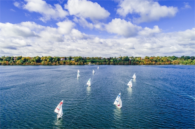 A group of small sailboats glides across a wide, blue lake under a partly cloudy sky, with a tree-lined shoreline and autumn foliage visible in the background.