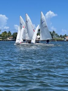 Three sailboats with white sails race on a sunny day, gliding across blue water with palm trees and houses visible along the shoreline under a bright blue sky with scattered clouds.