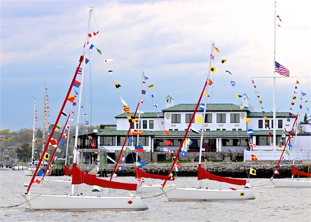 Three white sailboats with red sails and colorful signal flags float on the water near a waterfront building decorated with matching flags. The sky is cloudy, and an American flag flies on a pole nearby.