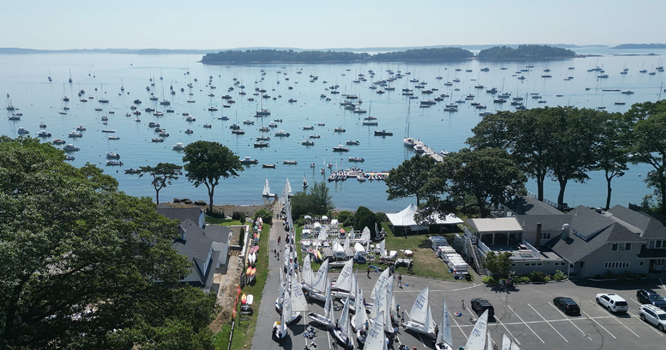 Aerial view of a coastal marina filled with many small sailboats on calm water, with islands in the distance and sailboats lined up on land near waterfront buildings and trees.