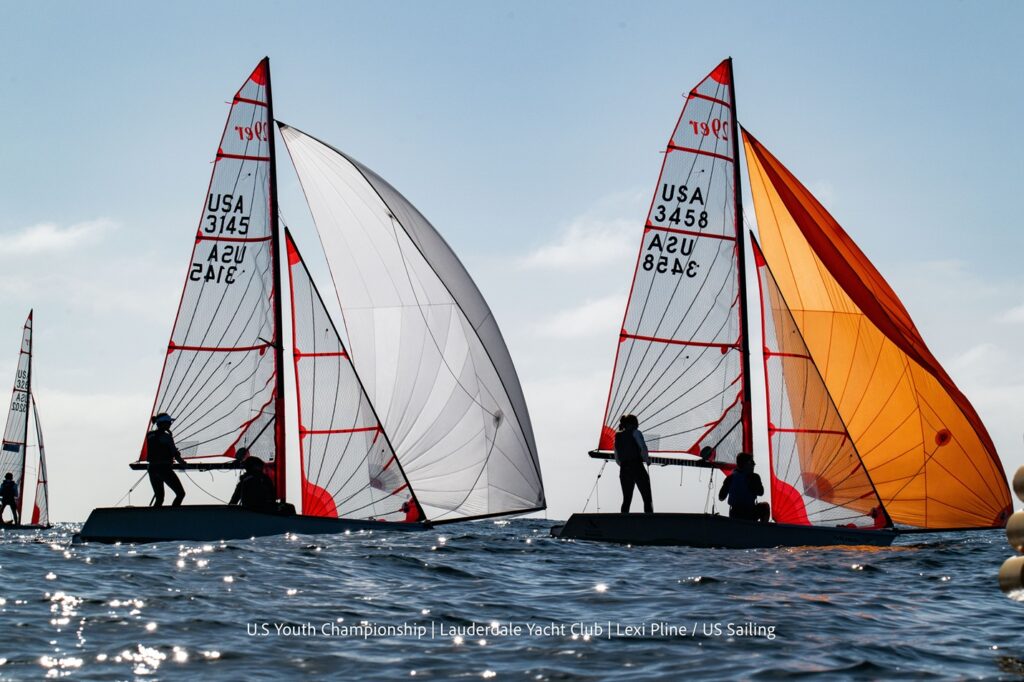 Two sailboats with white and orange sails, each with three sailors, race side by side on the water under a clear sky. Sail numbers and "USA" are visible on the sails. Other boats can be seen in the background.