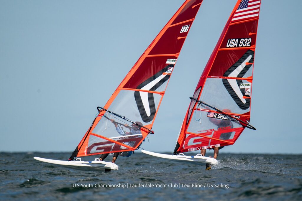 Two windsurfers race across the water on red sailboards, each with visible sail numbers and a US flag. The scene is set under a clear sky at the U.S. Youth Championship.
