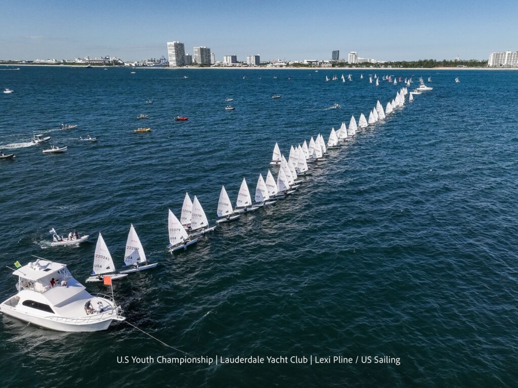 A long line of small sailboats races across blue water near a city shoreline, with a white motorboat at the front and many buildings visible in the background under a clear sky.