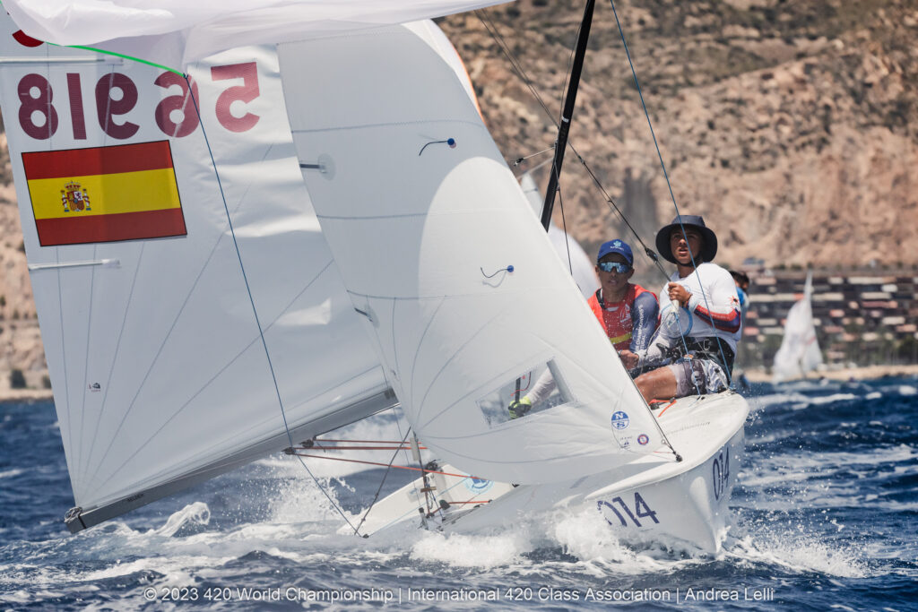 Two sailors maneuver a small sailboat with a Spanish flag on the sail during a race on the ocean, with a rocky coastline and buildings visible in the background.