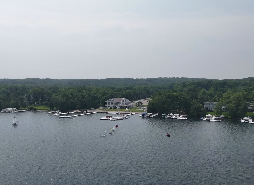 Aerial view of a lakeside marina with several docks, boats, and a large building surrounded by trees. A few small boats are on the water near the shore, and the background consists of dense forest under a cloudy sky.