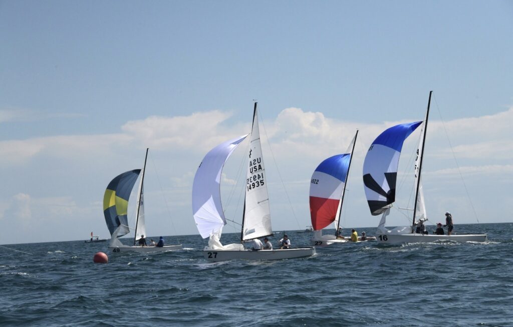 Four small sailboats with colorful spinnaker sails race on the open water beneath a blue sky with scattered clouds. The sea is slightly wavy, and a red buoy floats nearby.