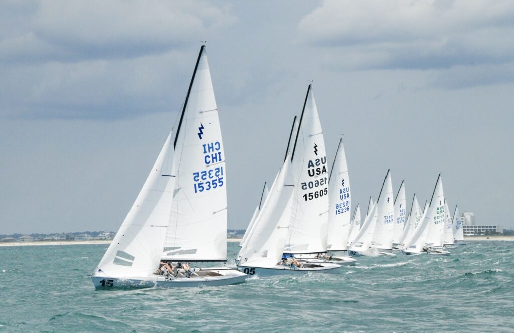 Several sailboats with white sails race in the ocean under a cloudy sky, with the distant shoreline visible on the horizon. Sailors are visible on each boat, and sail numbers are clearly marked.