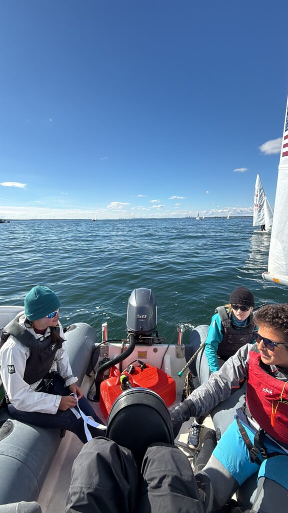 Three people in sailing gear sit in a small motorboat on calm blue water under a clear sky. One person steers while two others look on. Sailing boats are visible in the background.