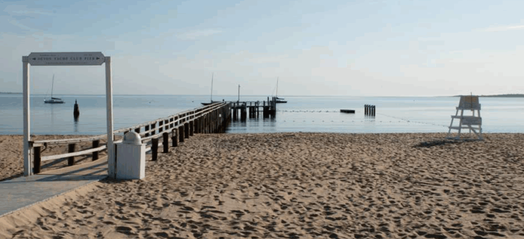 A sandy beach leads to a wooden pier extending over calm water, with a few boats anchored nearby. A lifeguard chair sits on the right, and the sky is clear and blue.